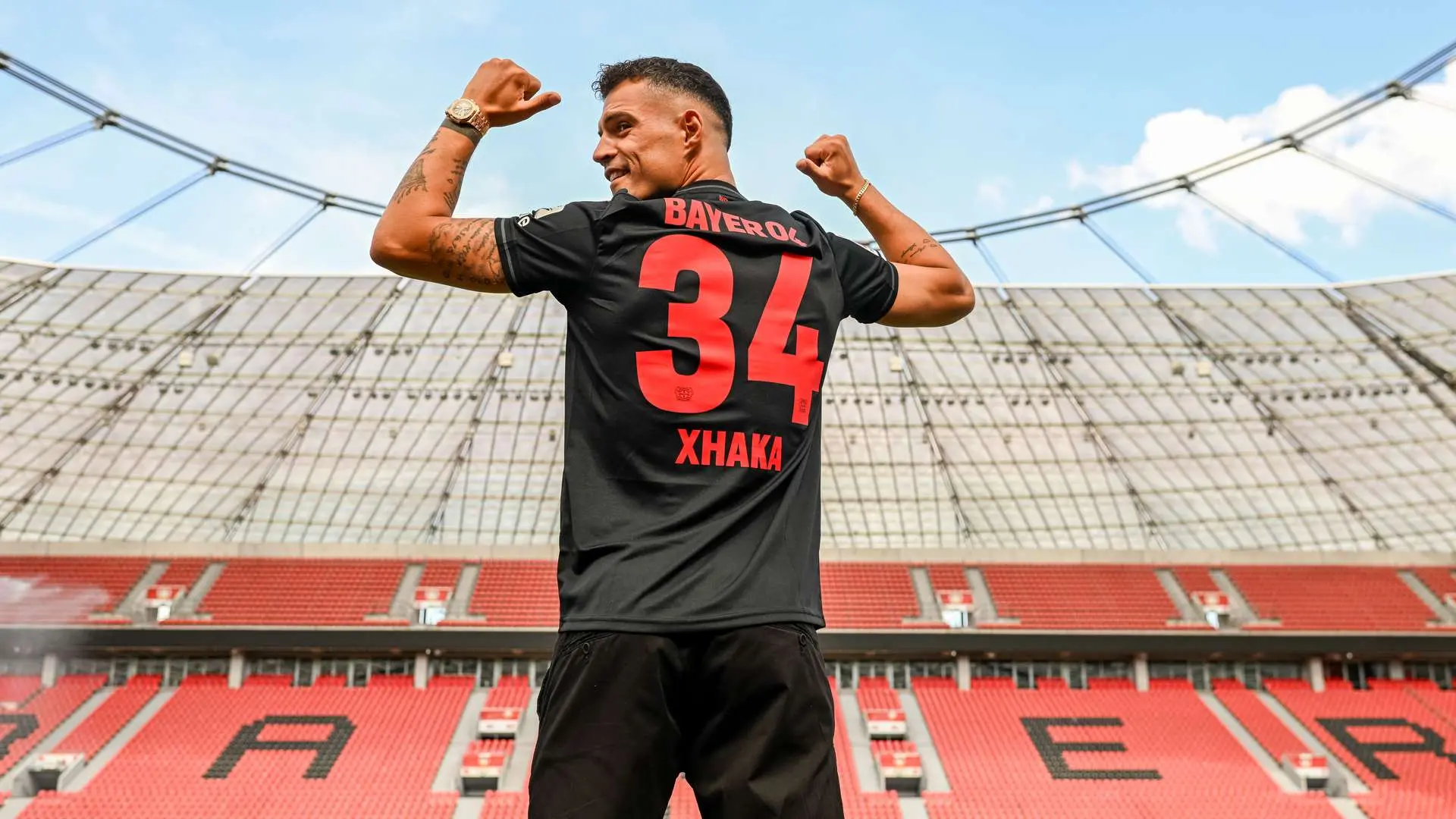 Granit Xhaka wearing a black Bayer Leverkusen jersey with number 34, celebrating in an empty stadium.