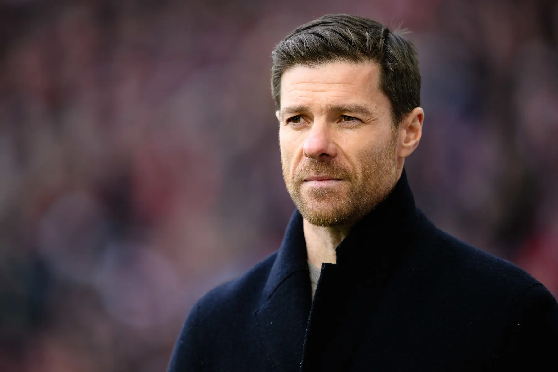 Close-up portrait of Xabi Alonso wearing a dark navy coat with a blurred stadium background.