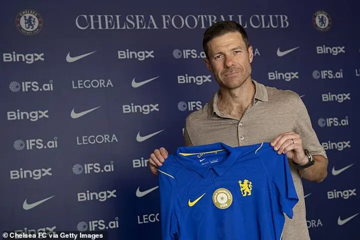 Xabi Alonso holding a Chelsea FC jersey in front of club branding.