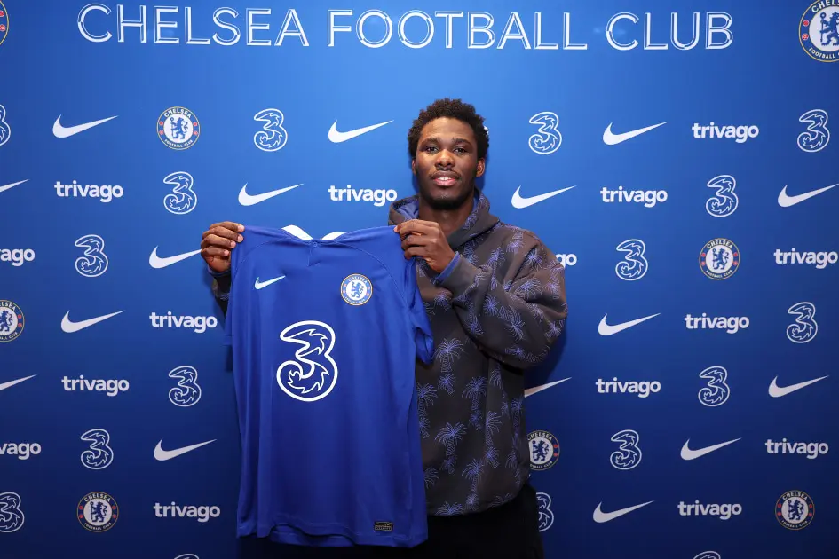 David Datro Fofana holding up a blue Chelsea FC home jersey in front of a branded club backdrop