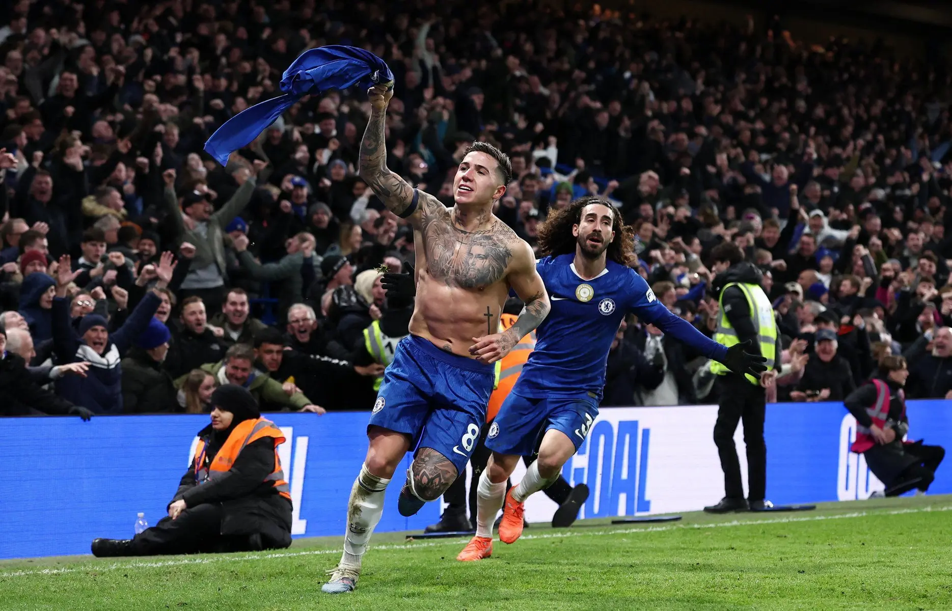 Enzo Fernandez celebrates shirtless with his jersey in hand, joined by a teammate during a Chelsea match.