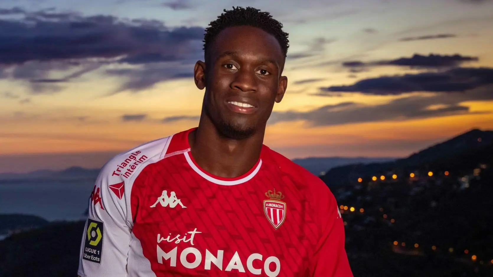 Folarin Balogun wearing the AS Monaco home kit, posing against a sunset landscape of Monaco.