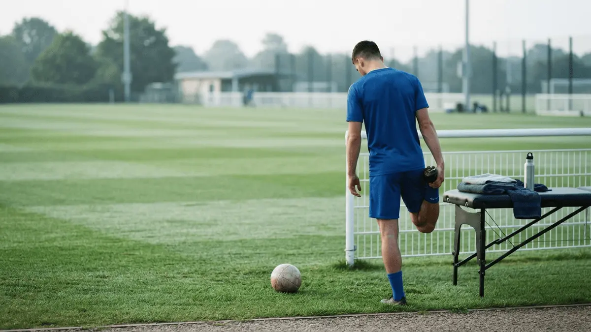 Non-identifiable footballer in royal blue training kit stretching a calf on a quiet training pitch