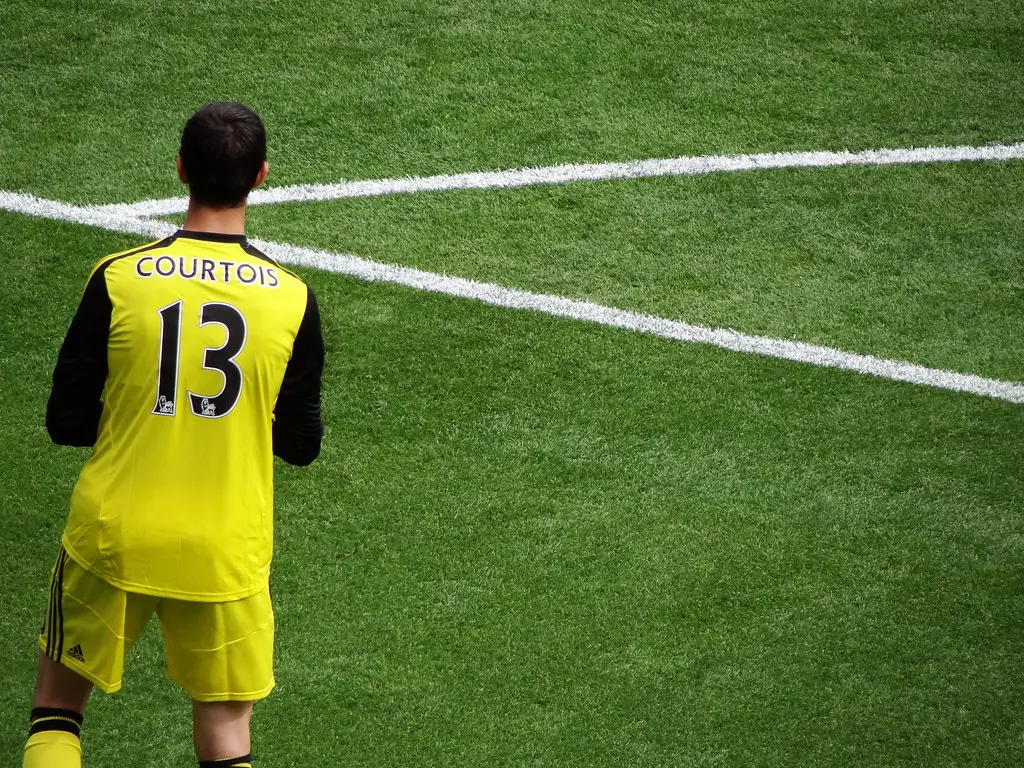 Thibaut Courtois seen from behind in a yellow Chelsea goalkeeper kit on a grass pitch.
