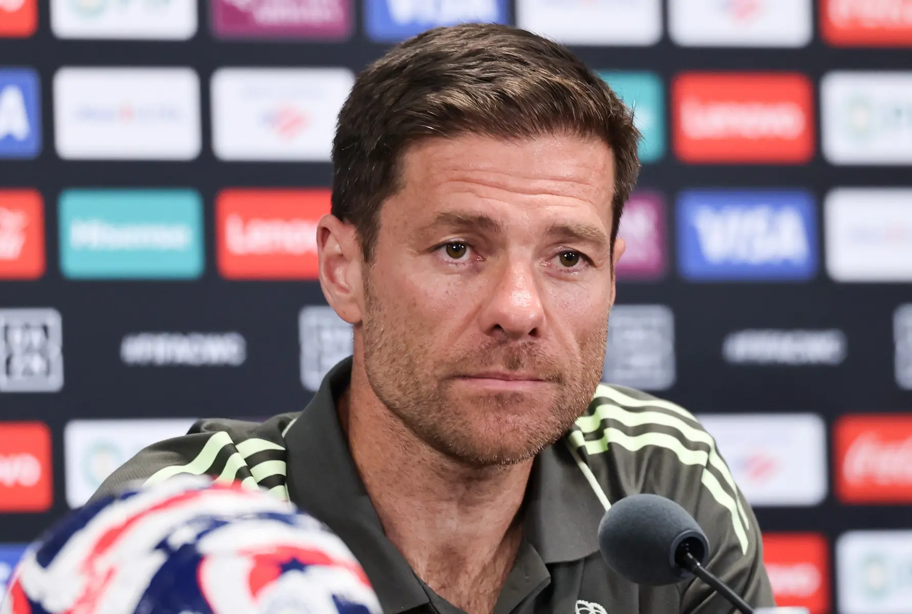 Xabi Alonso sitting at a press conference table with microphones and a branded sponsor backdrop.