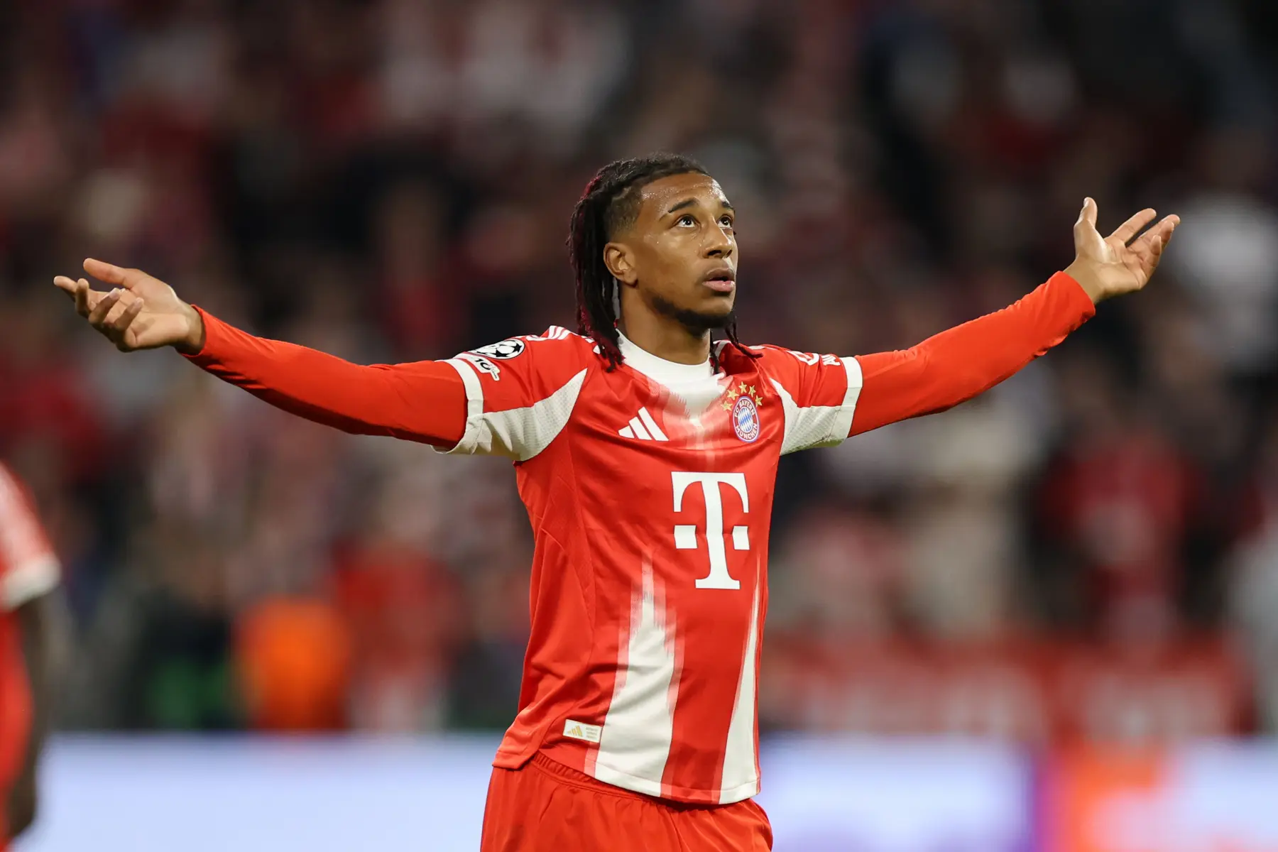 Michael Olise in a red Bayern Munich kit with arms outstretched during a match.
